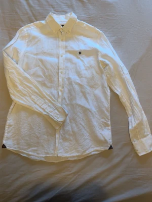 Vit linneskjorta från Morris - Klassisk vit skjorta från Morris i svalt linne. Skjortan har button-down krage, lång ärm och en liten broderad logga på bröstet. Perfekt för en clean och stilren look, lätt att matcha med jeans eller chinos.