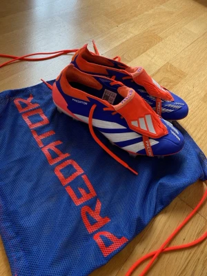 Adidas Predator elite fotbollskor - Säljer ett par Adidas Predator elite fotbollsskor i blått, rött och vitt. Dem är bara använda en gång och skorna har klassisk låg profil, snörning och mönstrad sula för bra grepp på planen. Tillverkade i syntetmaterial och levereras med en blå påse med röd Predator-logga. 