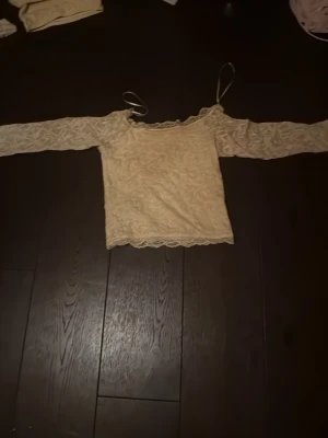 Beige spetsblus med långa ärmar - Säljer en söt beige blus i spets med långa ärmar och tunna axelband. Toppen har en offshoulder-design och fina spetsdetaljer längs kanterna. Perfekt för dig som gillar romantisk och feminin stil.