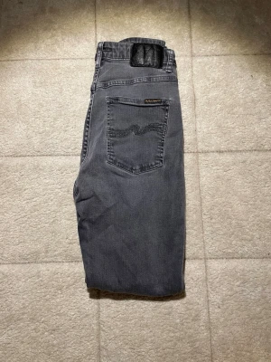 Grå jeans från Nudie Jeans - Snygga grå jeans från Nudie Jeans med klassisk femficksmodell och svarta detaljer på bakfickan. Jeansen har raka ben och är tillverkade i mjuk bomull. Perfekta för en avslappnad och stilren look.