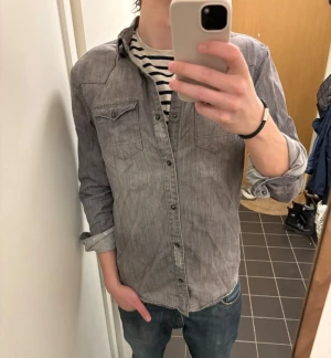 Grå skjorta från AllSaints - Säljer en grå långärmad skjorta från AllSaints med knappar framtill och två bröstfickor med lock. Skjortan har en avslappnad passform och är tillverkad i mjuk bomull. Perfekt att bära öppen över en t-shirt eller stängd för en clean look.