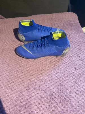  Nike Mercurial fotbollsskor - Säljer ett par blå Nike Mercurial fotbollsskor med högt skaft och vita detaljer. Skorna har en strumpliknande passform och är tillverkade i ett lätt syntetmaterial för bra bollkontroll. Insidan är gul och yttersulan har dobbar för spel på gräs.
