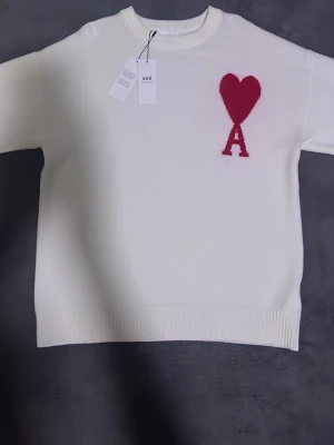 Vit stickad tröja  - Vit stickad tröja  med röd hjärt- och A-detalj på bröstet. Tröjan har rund halsringning, ribbade muddar och en avslappnad passform. Perfekt för dig som gillar stilrena plagg med en twist. Fråga gärna om fler bilder 