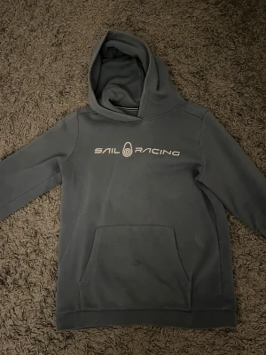 Sail Racing hoodie - Mörkgrå hoodie från Sail Racing med stor magficka och vit logga tryckt över bröstet. Klassisk huva och avslappnad passform, perfekt för chill dagar. Tillverkad i mjukt material som känns skönt mot huden.