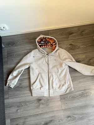  Burberry Hoodie  - Säljer en grå zip-hoodie från Burberry med klassiskt rutigt foder i huvan. Hoodien har två fickor fram, dragsko i huvan och ribbade muddar. Perfekt för dig som gillar stilrena plagg med en twist. Materialet är mjukt och bekvämt bomullstyg.