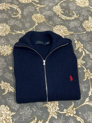 Marinblå ziptröja från Polo Ralph Lauren - Snygg marinblå stickad ziptröja från Polo Ralph Lauren med ribbad struktur och röd broderad logga på bröstet. Tröjan har hög krage, dragkedja framtill och breda muddar vid ärmslut och nederkant. Perfekt för lager på lager och stilren vardagslook.