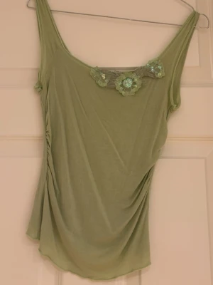 Grön linnetopp med spetsdetalj - Snygg ljusgrön linnetopp med smala axelband och dekorativa spetsblommor med pärlor vid urringningen. Toppen har draperad effekt på sidan och är tillverkad i ett mjukt, tunt material som faller fint.