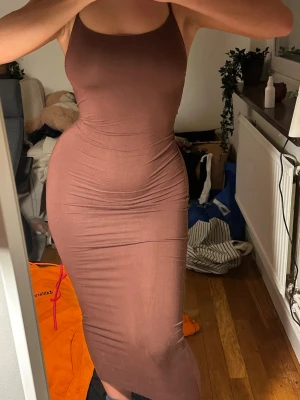 Brun tight långklänning med smala band - Säljer en brun långklänning med smala axelband som korsas i ryggen. Klänningen är figurnära och har öppen rygg med en snygg rynkdetalj baktill. Perfekt för dig som gillar en stilren och trendig look.