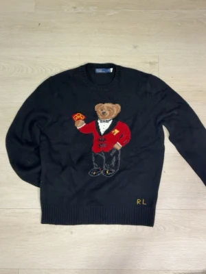Svart stickad tröja med björnmotiv RL - Stickad svart tröja från Ralph Lauren med ikoniskt björnmotiv på bröstet. Björnen har röd kavaj och håller en accessoar. RL-broderi i gult vid nederkanten. Rund halsringning och ribbade muddar. Perfekt för dig som gillar statement-plagg.