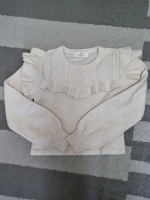 Beige stickad tröja med volang - Supersöt beige stickad tröja från Lindex med volangdetaljer över axlarna och ett diskret mönster framtill. Tröjan har rund halsringning, långa ärmar och ribbade muddar. Perfekt för dig som gillar en romantisk och trendig look.