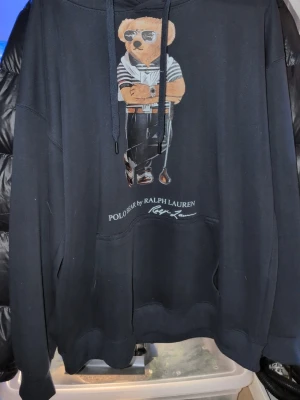 Polo Bear hoodie - Mörkblå hoodie från Ralph Lauren med Polo Bear-tryck framtill. Klassisk huva med dragsko och stor känguruficka. Mjuk bomullsblandning och avslappnad passform. Perfekt för en cool och avslappnad stil. Säljer min polo bear hoddie desperat efter pengar😅 står strl xxl men den är absolut inte ens nära de den sitter typ exakt som en normal L. Qr kod scannern funkar och man kommer in på deras hemsida vet inte till 100 om den är the real deal. Riktigt skönt och extremt snygg till golfen i sommar😉0skador