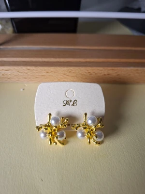 Guldörhängen med pärldetaljer - Snygga örhängen i guldfärgad metall med vita pärlor. Designen är inspirerad av blommor med tre pärlor i varje örhänge och organiska guldfärgade detaljer runt om. Perfekta för att ge din look en elegant touch.