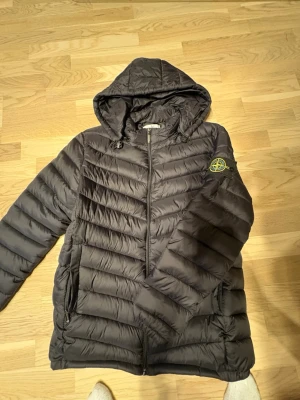 Svart pufferjacka från Stone Island - Säljer en svart pufferjacka från Stone Island med huva och dragkedja framtill. Jackan har det klassiska Stone Island-märket på vänster ärm och två sidofickor med dragkedja. Tillverkad i ett glansigt, quiltat material som håller värmen under kalla dagar.