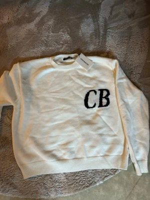 Vit stickad tröja CB Cole Buxton  - Vit stickad tröja från Cole Buxton med svarta bokstäver 'CB' på bröstet. Tröjan har rund halsringning, ribbade muddar och är tillverkad i mjukt material. Enkel och stilren design som passar till mycket.