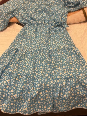 Blommig blå kortklänning med volang - Söt blå kortklänning med vitt och gult blommönster. Klänningen har volangdetaljer, v-ringning och resår i midjan för en bekväm passform. Perfekt för vår och sommar, med en luftig och lätt känsla.