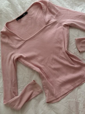 Rosa ribbad topp med v-ringning - Snygg ljusrosa ribbad topp från Bik Bok med lång ärm och v-ringad hals. Figurnära passform och mjukt, stretchigt material som sitter skönt på kroppen. Perfekt att styla med jeans eller kjol för en trendig look.