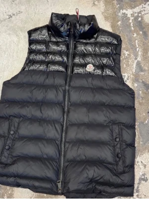 Svart dunväst från Moncler - Snygg svart dunväst från Moncler med hög krage och dragkedja framtill. Västen har två fickor med tryckknappar och det klassiska Moncler-logotypen på bröstet. Tillverkad i glansigt material som ger en exklusiv känsla.