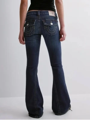 Mörkblå bootcut jeans True Religion - Säljer ett par mörkblå bootcut jeans från True Religion med ikoniska kontrastsömmar och stora fickor bak med vita knappar. Jeansen har låg midja och klassisk femficksdesign. Perfekta för dig som gillar en retro och avslappnad stil.
