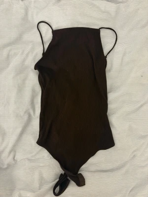 Brun ribbad body med smala axelband - Snygg brun body med ribbad struktur och smala axelband. Modellen har rundad halsringning och tryckknappar i grenen för enkel på- och avtagning. Perfekt att styla till jeans eller kjol. Materialet känns mjukt och stretchigt.