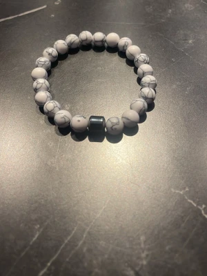 Elastiskt armband med vita pärlor - Snyggt armband med vita, marmorerade pärlor och en större mörkgrå pärla i mitten. Armbandet är elastiskt och passar de flesta handleder. Perfekt accessoar för att lyfta din stil med en clean och modern touch.