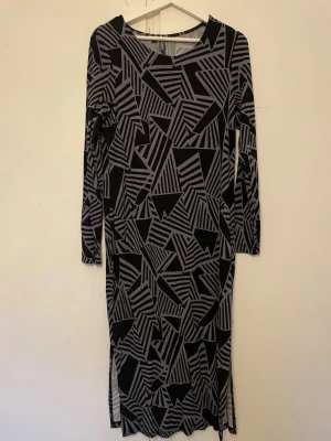Svart och grå långklänning med mönster - Snygg långklänning i svart och grått med grafiskt mönster. Klänningen har långa ärmar och en rak passform. Tillverkad i mjukt material som känns bekvämt mot huden. Perfekt för dig som gillar unika prints och stilrena färger.