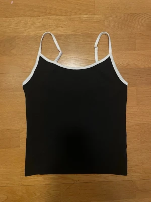 Svart linnetopp med vita kanter - Snygg svart linnetopp med tunna, justerbara axelband och vita kontrastkanter. Enkel och stilren design som passar perfekt till jeans eller kjol. Mjuk och stretchig känsla för en bekväm passform.