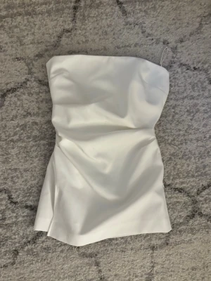 Vit axelbandslös topp från Zara - Snygg vit bandeautopp från Zara. Toppen har en enkel och stilren design med figurnära passform och är tillverkad i ett slätt material. Har några väldigt små fläckar (se bild 5), men de syns inte då de ligger som ”mellan lagren” 💕