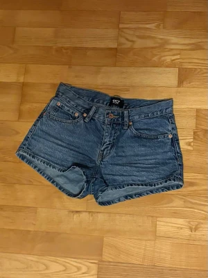 Blå jeansshorts från lager 157 - Säljer ett par klassiska blå jeansshorts från lager 157 med fem fickor, bälteshällor och knappgylf. Shortsen har en avslappnad passform och är tillverkade i slitstarkt denimtyg. Perfekta för sommaren och enkla att matcha med olika stilar.