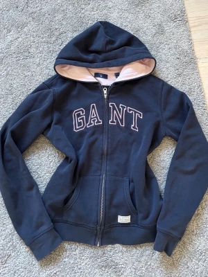 Marinblå hoodie med dragkedja GANT - Mysig marinblå hoodie från GANT med dragkedja framtill och stor logga i vitt och rosa på bröstet. Tröjan har huva med ljusrosa foder och två fickor fram. Perfekt för en avslappnad och sportig stil. Storlek 158-164 skulle säga ca Xs