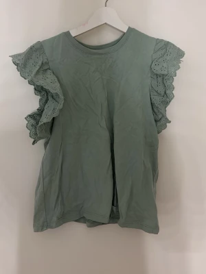 Grön topp med volangärmar - Snygg grön topp med dubbla volangärmar i broderad spets. Toppen har rund halsringning och är tillverkad i mjuk bomull. Perfekt för dig som vill ha en söt och trendig look med extra detaljer på ärmarna.