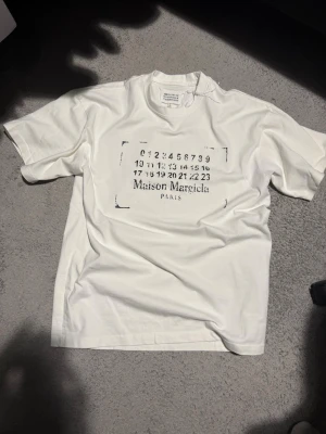 Maison Margiela vit t-shirt med tryck - Snygg vit t-shirt från Maison Margiela med ikoniskt svart tryck framtill. Klassisk passform och rund hals. Perfekt för dig som gillar minimalistisk och stilren design. Tillverkad i mjuk bomull för skön känsla hela dagen.