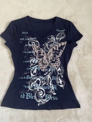 Mörkblå t-shirt med fjärilstryck - Snygg mörkblå t-shirt med v-ringning och ett stort tryck av en fjäril och vita mönster på framsidan. T-shirten har korta ärmar och är figurnära i modellen. Perfekt för dig som vill sticka ut lite extra.