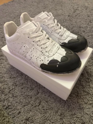 Maison Margiela svartvita sneakers - Unika sneakers från Maison Margiela i vitt läder med svarta detaljer och färgstänk. Lågt skaft och klassisk siluett med vita skosnören och svartmålade tåpartier. Perfekta för dig som gillar statement-plagg och vill sticka ut med din stil.