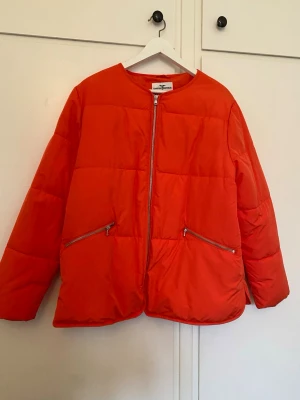 Orange pufferjacka från Hampton Republic - Snygg orange pufferjacka från Hampton Republic med raka linjer och två snedställda fickor med dragkedja. Jackan har en enkel design med rund halsringning och grov dragkedja framtill. Perfekt gör våren och jackan äf boxig och oversize. Ser röd ut på bilden men är orange. 