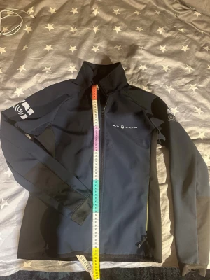 Marinblå vindjacka från Sail Racing - Snygg marinblå vindjacka från Sail Racing med hög krage och dragkedja framtill. Jackan har logga på bröstet och armen, samt diskreta svarta detaljer längs sidorna och ärmarna. Tillverkad i ett vindtätt material som passar perfekt för blåsiga dagar.