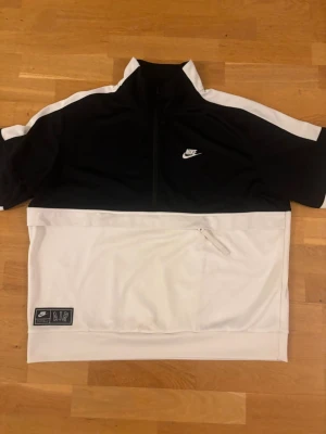 Nike svartvit hoodie med dragkedja - Snygg svartvit hoodie från Nike med halv dragkedja framtill och vit logga på bröstet. Hoodien har vita detaljer längs ärmarna och en stor ficka med dragkedja på magen. Tillverkad i mjukt och bekvämt material, perfekt för en sportig och avslappnad stil.