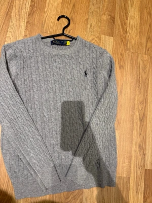 Grå kabelstickad tröja Polo Ralph Lauren - Kabelstickad grå tröja från Polo Ralph Lauren med rund halsringning och diskret broderad logotyp på bröstet. Tröjan har långa ärmar och ribbade muddar vid ärmslut och nederkant. Perfekt för en stilren och klassisk look.