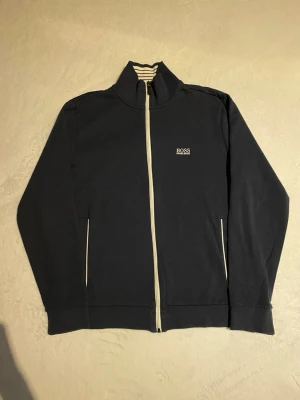 Svart ziptröja från Hugo Boss - Snygg svart ziptröja från Hugo Boss med vit dragkedja och broderad logga på bröstet. Tröjan har ståkrage, lå sopassform och två fickor framtill. Insidan av kragen har randigt tyg i vitt och svart. navy spectrum super clean.