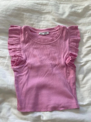 Rosa ribbad topp med volangärm Zara - Supersöt rosa topp från Zara i ribbat material. Modellen har korta ärmar med dubbla volangdetaljer som ger en trendig och lekfull look. Rund halsringning och figurnära passform. Perfekt att styla till jeans eller kjol för en fräsch outfit.