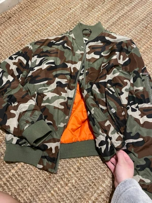 Camo bomberjacka från Junkyard XS - Säljer en camouflagemönstrad bomberjacka från Junkyard i storlek XS. Jackan har klassisk grön, brun och beige camo, orange innerfoder och ribbade muddar vid ärmar och midja. Två snedställda fickor framtill och ärmficka med dragkedja. Perfekt statement-plagg!
