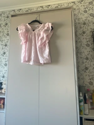 Ljusrosa volangblus med smock - Supersöt ljusrosa blus med volangdetaljer vid ärmhålen och smockad halsringning. Blusen är ärmlös och har en luftig, avslappnad passform. Perfekt för en trendig och feminin look. Materialet ser ut att vara lätt bomull eller linne.