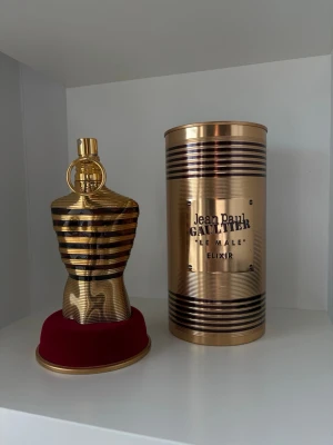Jean Paul Gaultier Le Male Elixir - Lyxig parfym i guldigt, metalliskt flakong formad som en manlig torso med röda ränder. Kommer med en matchande guldig cylinderformad förpackning med texten 'Le Male Elixir'. Perfekt för dig som gillar exklusiva dofter och snygg design. Har länge varit en parfymfantast och älskar parfymer men har på senaste tappat suget lite, har därför förvarat alla mina parfymer i mörker och under goda förhållanden. (Du ser hur mycket som är kvar på tredje bilden)