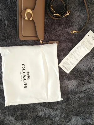 Coach Tabby 26 Bag i brunt läder - Säljer en snygg Coach Tabby Bag i brunt pebble-läder med guldfärgade detaljer och avtagbar axelrem. Klassisk rektangulär form och elegant spänne framtill. Perfekt för dig som vill ha en stilren och trendig handväska från Coach.