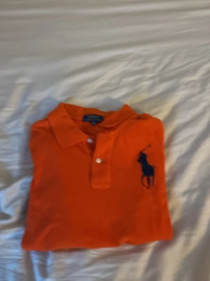 Orange pikétröja från Ralph Lauren - Snygg orange pikétröja från Ralph Lauren med klassisk krage och två knappar framtill. Stor mörkblå logga broderad på bröstet. Perfekt till jeans eller shorts för en avslappnad men stilren look.är långarmmad