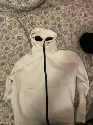 Vit hoodie med glasögon från C.P. Company - Säljer en vit hoodie från C.P. Company med hel dragkedja och ikoniska inbyggda glasögon i huvan. Hoodien har broderad logga på bröstet. Riktigt skön hoodie för bra pris 