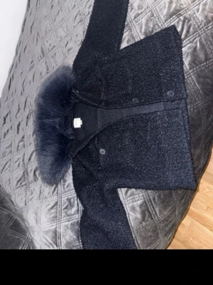  pälsjacka i mörk blå - Säljer en mörkblå pälsjacka med fluffig fuskpälskrage. Jackan har knappar framtill och en kort, boxig passform. Materialet är mjukt och lurvigt, perfekt för kalla dagar. Kragen ger en extra lyxig känsla och jackan är riktigt snygg till vintern och vår.