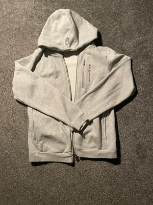 Grå hoodie från Sail Racing - Säljer en grå hoodie från Sail Racing med dragkedja och huva. Hoodien har ett broderat Sail Racing-logo på bröstet och en praktisk bröstficka med dragkedja. Insidan är mjuk och fleecefodrad för extra komfort. Perfekt för dig som gillar stilren design. Var för stor för mig 