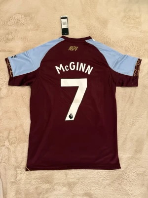 Aston Villa matchtröja McGinn #7 - Snygg Aston Villa matchtröja från Adidas med McGinn och nummer 7 på ryggen. Tröjan är vinröd med ljusblå ärmar, vita detaljer och klubbmärke på bröstet. Tillverkad i lätt och ventilerande material, perfekt för fotboll eller träning.