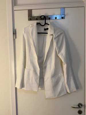 Vit kavaj med enkel knapp - Stilren vit kavaj med klassisk passform och en knapp framtill. Kavajen har två fickor och är helfodrad. Perfekt för en clean och fräsch look. Materialet ser ut att vara bomull eller en bomullsblandning. Inga synliga detaljer från något märke. Ett sett med kostym byxor och kavaj. Använd endast en gång till studenten 