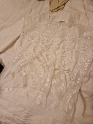 Vit spetsblus från Kaffe - Säljer en superfin vit spetsblus från Kaffe i storlek M. Blusen har transparent mesh med broderade blommor och blad, rund halsringning och korta ärmar. Perfekt för dig som gillar romantiska detaljer och en luftig känsla.